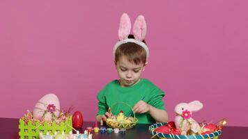 contento pequeño niño arreglando huevos y polluelo en un cesta en preparación para Pascua de Resurrección domingo celebraciones, creando festivo decoraciones en estudio. pequeño chico teniendo divertido colorante. cámara una. foto