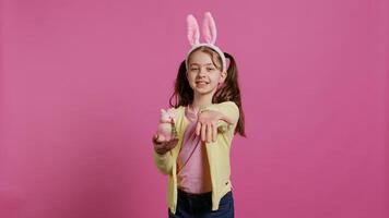 Lovely little girl with bunny ears sending air kisses in studio, presenting a stuffed pink rabbit toy for easter holiday. Smiling energetic preteen feeling positive and carefree. Camera B. photo