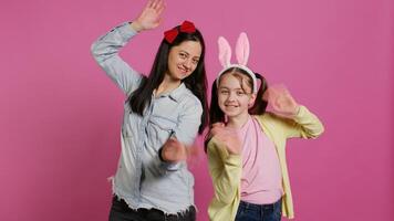 sonriente alegre madre y pequeño niña ondulación en frente de cámara, teniendo divertido y disfrutando Pascua de Resurrección fiesta celebracion. alegre Chica de escuela posando con su mamá en estudio, diciendo Hola. cámara b. foto