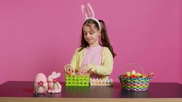 alegre pequeño niña demostración su hecho a mano festivo cesta en cámara, creando un vistoso hecho a mano arreglo en hora para Pascua de Resurrección día festivo. pequeño niñito con conejito orejas decorando huevos. cámara b. foto