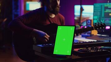 Composer performing acoustical guitar in his home studio, playing and practicing on strings. Musician learning multiple songs on instrument, uses tablet to run greenscreen display. Camera A. photo