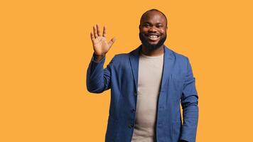 African american man cheerfully doing salutation hand gesture. Portrait of happy BIPOC person raising arm to greet someone, gesturing, isolated over yellow studio background, camera A photo