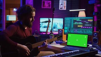 Musician beginner learning accords to play acoustic guitar by watching tutorials and online lessons, laptop shows greenscreen chromakey. Artist practicing guitar skills in home studio. Camera B. photo