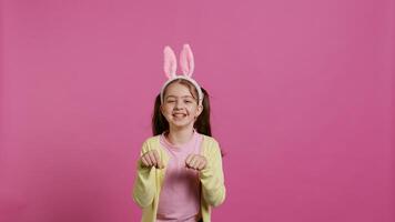 Joyful carefree schoolgirl jumping around in studio, imitating a rabbit and hopping against pink background. Cheerful active child wearing bunny ears and bouncing, adorable kid. Camera B. photo