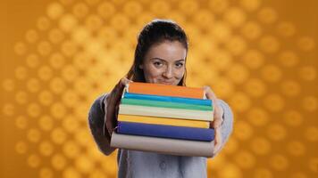 Portrait of friendly woman offering stack of textbooks useful for school exam, isolated over studio background. Merry person giving pile of books, recommending them for university assessment, camera B photo