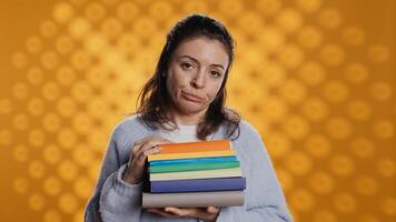 Portrait of woman with pouting expression holding pile of books, showing disproval of reading hobby. Sulky lady with stack of novels doing thumbs down hand gesturing, studio background, camera B photo