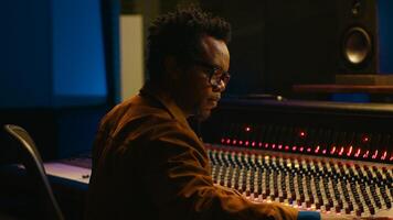 Portrait of african american audio technician uses mixing console with knobs and sliders in control room, editing recordings and adding sound effects. Skilled music producer at mixer. Camera A. photo