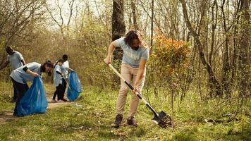 Young man covering base and hole after planting small tree in the woods, working with shovel to grow seedlings and preserve forest habitat. Activist does voluntary work for the planet. Camera B. photo