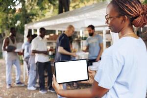 An isolated mockup template for philanthropic messages is held by young black woman holding a digital tablet. African american woman volunteer holding a smart device with a blank white screen display. photo