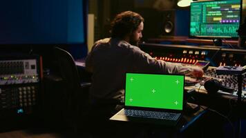 Music production engineer mixing and mastering music in control room, using laptop with greenscreen display. Skilled audio technician working with recording software on pc, pushing faders. Camera B. photo