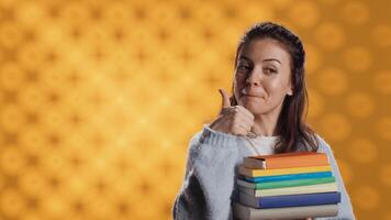 retrato de contento mujer con apilar de libros en manos demostración pulgares arriba, estudio antecedentes. jubiloso ratón de biblioteca participación pila de novelas, sensación optimista, haciendo positivo mano gesticulando, cámara un foto