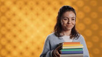 Portrait of smiling woman holding stack of books, doing salutation hand gesture, feeling optimistic. Jolly person with pile of novels raising arm to greet someone, studio background, camera A photo