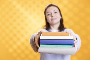Portrait of smiling woman offering pile of books, recommending reading hobby for relaxation purposes. Radiant bookaholic person with stack of novels in arms doing endorsement, studio background photo