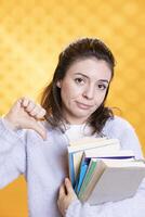 Portrait of woman holding stack of books doing thumbs down sign gesturing, studio background. Stressed student with pile of textbooks in arms used for academic learning doing negative hand gesture photo