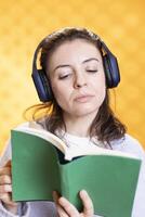 Woman concentrating on reading book, relaxing and listening audio white noise, isolated over studio background. Geek with novel in hand hearing songs through headphones, entertainment concept photo