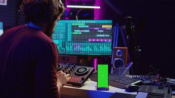 Sound engineer working on mixing and mastering techniques in his home studio, using phone with greenscreen display while he is recording audio. Artist operating soundboard and amplifier. Camera B. photo