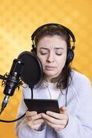 Narrator frowning while reading aloud from ebook on ereader into professional mi against backdrop, doing dramatic acting. Woman recording audiobook, reading text from tablet, portraying characters photo