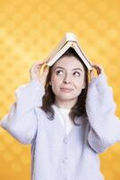 Portrait of joyous woman placing book on head, acting zany, enjoying reading hobby. Upbeat lady having fun wearing novel as hat, clowning around, isolated over studio background photo