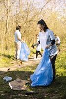 Female volunteer tidying the woodland of garbage and plastic bottles, collecting rubbish with claw and disposal bag. Young activist volunteering to conserve natural ecosystem. photo