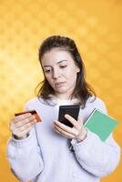 Woman appalled by books cost while adding credit card information on cellphone, studio background. Person balking at high price of academic textbook needed for university classes, using phone photo