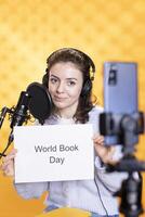 Woman filming promotional for world book day using microphone and phone, studio background. Content creator promoting reading, gaining awareness for literature importance photo