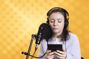 Narrator wearing headphones reading aloud from ebook on ereader into professional microphone against backdrop. Voice actor recording audiobook, reading text from tablet screen, doing audio story photo