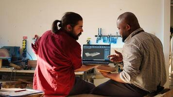 African american manufacturer and colleague comparing wood art piece with schematic displayed on CAD software on laptop. Woodworking professionals crosschecking wooden object with blueprint, camera B photo