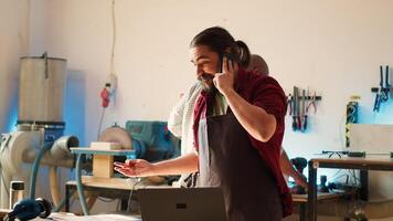 Manufacturer at work receiving telephone call from client inquiring about commission progress. Carpenter in assembly shop having friendly conversation on cellphone with customer, camera A photo