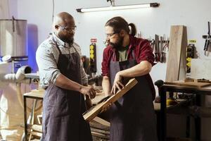 Carpenter holding timber block, brainstorming with coworker next steps in wood processing. Manufacturer in joinery and african american apprentice discussing what to do with piece of wood photo