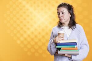 Portrait of woman with stack of books in hands holding disposable cup of coffee, isolated over studio background. Bookworm holding pile of novels and caffeinated drink early in the morning photo