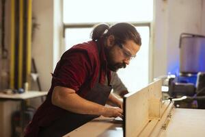 Man wearing protection equipment while working with spindle moulder to prevent workplace accidents. Cabinetmaker equipped with safety goggles while cutting with wood shaper to avoid injury photo