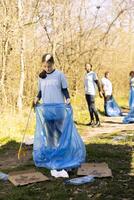 Community service volunteer reduces waste and cleans up forest setting, collecting garbage in blue trash bags. Young kid helps environment conservation by disposing of plastic waste. photo