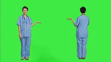 Medical assistant pointing at something aside and doing web commercial, creating advertisement against greenscreen backdrop. Young nurse with scrubs showing a thing to left or right sides. Camera A. photo