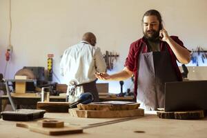 Smiling manufacturer discussing on telephone call with client inquiring about furniture assembling commission progress. Cheerful carpenter in assembly shop talking on mobile phone with customer photo