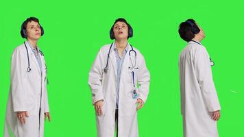 Joyful physician dancing around in the studio with music on headset, having fun with modern cool songs against greenscreen backdrop. Medic woman enjoying audio tunes and singing. Camera B. photo