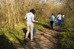 Young nature activist collecting junk with a long claw tool and garbage bag, grabbing plastic waste and trash to clean the woodland. Male volunteer does community service, global pollution. photo