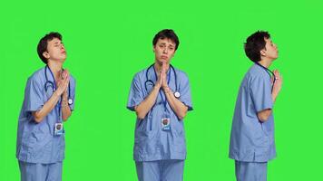 Medical assistant being hopeful praying for good luck against greenscreen, holds hands in a prayer sign and having belief in spirituality. Young christian nurse with faith in meditation. Camera B. photo