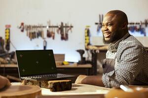 alegre carpintero y bipoc compañero de trabajo utilizando canalla software en verde pantalla ordenador portátil a diseño de madera objetos. alegre carpinteros utilizando programa en Bosquejo cuaderno a plan mueble montaje en carpintería foto