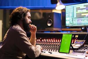 Skilled sound engineer looks at mockup display on tablet before mixing and mastering tracks on electronic stereo console. Technician uses digital audio software to record and edit his songs. photo