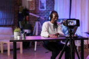 Focus shot on modern camera recording vlogger in blurry background talking about family and relationships subjects. Woman producing online show using high quality equipment in rgb lights studio photo