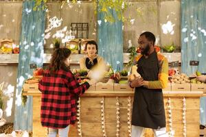 Black male farmer showing natural organic seasonal produce to customer at greenmarket kiosk. Group of vendors and clients at harvest fair festival, browsing freshly harvested fruits and vegetables. photo