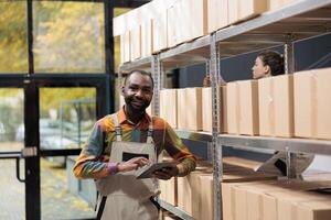 African american manager working at goods inventory, checking products on tablet computer in warehouse. Stockroom employee preparing customers packages, using cardboard boxes for delivery photo