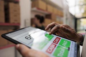 Worker checking online customers orders using tablet computer, preparing cardboard boxes before shipping. African american manager working at products inventory in warehouse. Close up concept photo