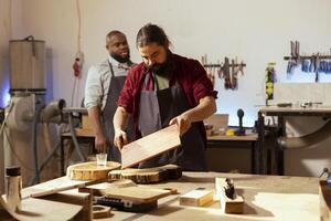 Craftsperson at workbench starting restoration project on wood block next to african american coworker. Carpenter preparing to color plank with paintbrush near apprentice solving tasks behind photo