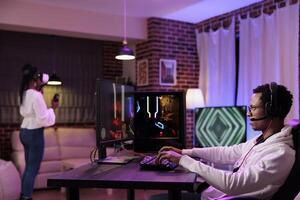 Gamer immersed in gaming session at computer desk while girlfriend in background uses virtual reality headset. African american couple playing games on gaming pc and vr system in rgb lights home photo