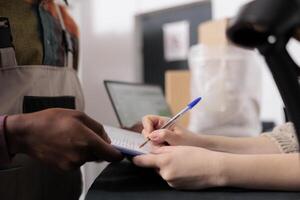 Storage room supervisor signing delivery documents, african american employee working at customers orders preparing packages in storehouse. Diverse team standing at counter in warehouse. Close up photo