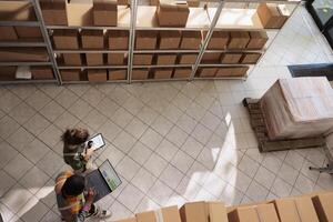 Diverse storage room workers tracking customers orders, checking warehouse logistics using laptop computer. Supervisor preparing packages for delivery with storehouse manager. Top view concept photo