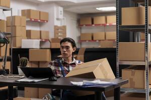 almacén empleado en silla de ruedas Procesando orden en ordenador portátil mientras participación cartulina caja Listo para envío. almacén asiático joven hombre entrega gerente trabajando en invalidez simpático lugar de trabajo foto