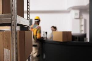 Selective focus of metallic shelves full with carton boxes, in background diverse employees discussing goods inventory report. Warehouse team working at customers orders, preparing packages photo