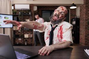 Weary man on office desk chair yelling in despair, feeling like being worked to death, looking like corpse. Lifeless businessman turned into zombie screaming in frustration from capitalism pressure photo